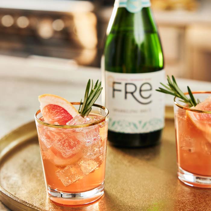 Orange cocktail with herb sprig garnish in front of a bottle of FRE Alcohol-Removed Sparkling Brut