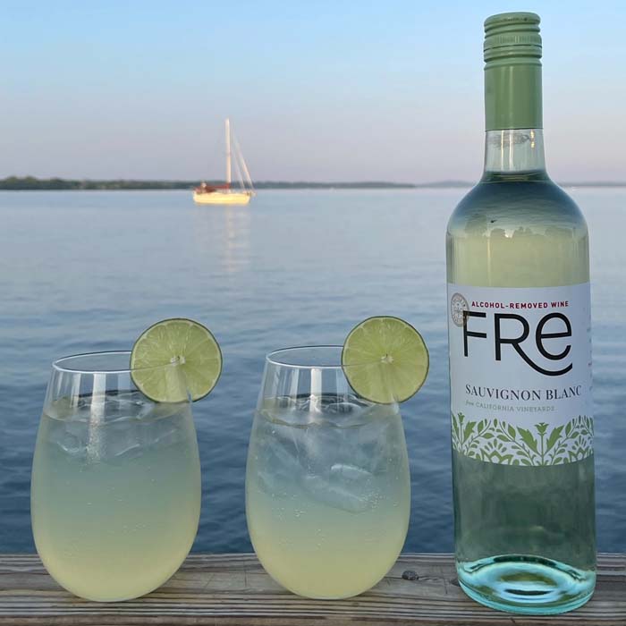 color image at sunset on the beach with a bottle of FRE Alcohol-Removed Sauvignon Blanc and 2 cocktails garnished with lime