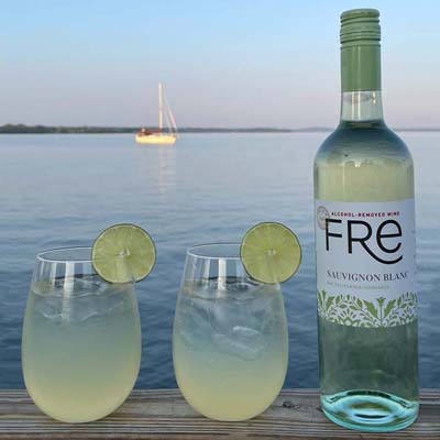 color image at sunset on the beach with a bottle of FRE Alcohol-Removed Sauvignon Blanc and 2 cocktails garnished with lime