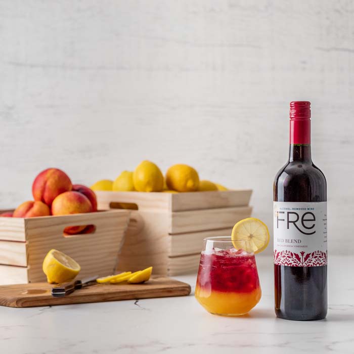 Color image of a bottle of FRE Alcohol-Removed Red Blend on a white counter next to a red and yellow cocktail with fruit in the background