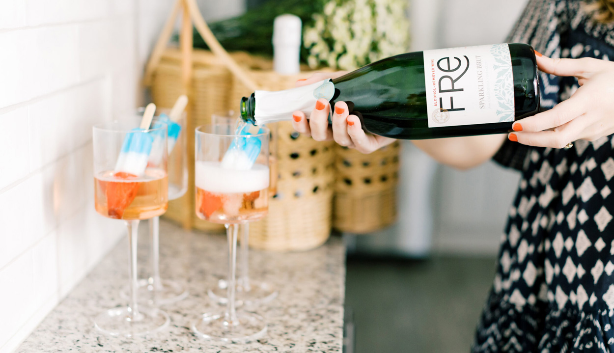 A woman's torso with her hands pouring a bottle of FRE Alcohol-Removed Sparkling Brut into two cocktail glasses on a marble surface containing liquid and red white and blue frozen pops, with wicker baskets and greenery in the background