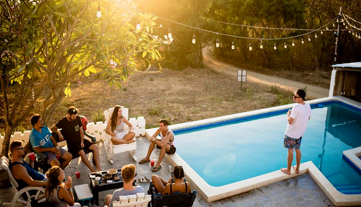 A summer scene of a group of seated people gathered on a patio next to a small swimming pool, with string lights overhead and a magnolia tree over the patio and woods in the background
